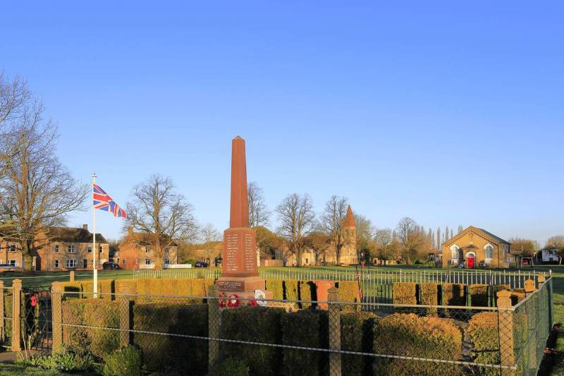 War memorial granite obelisk in Whittlesey with local buildings and church in background, sunny day, commemorating fallen heroes.