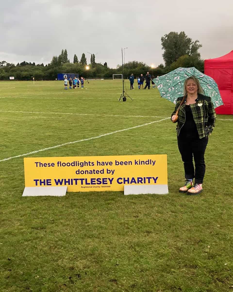 Floodlamp donation, The Whittlesey Charity, outdoor sports field, community support, charitable event, raincoat, woman with umbrella, charity fundraiser, donation sign, local charity support.